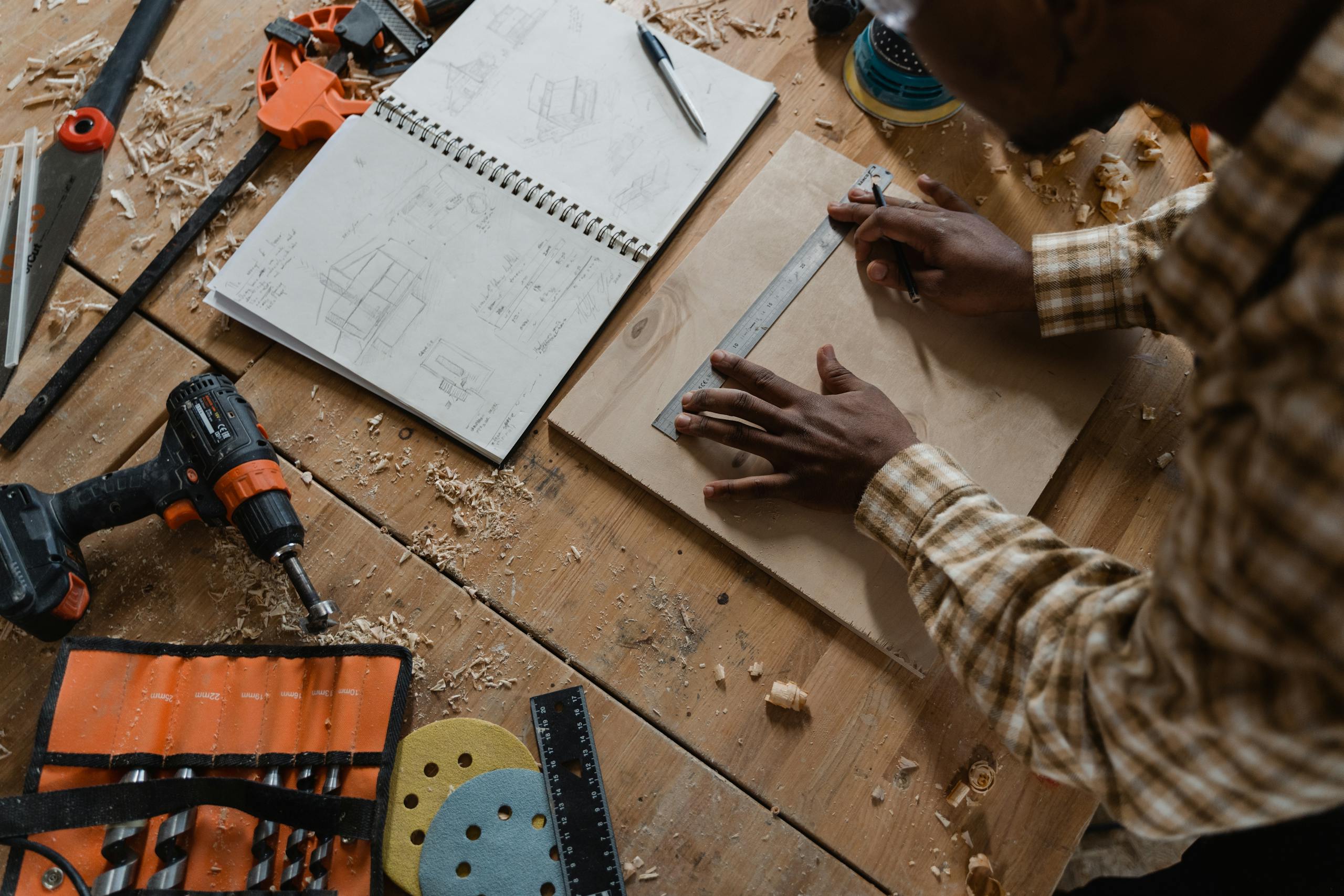 Woodworker measures timber amidst tools and sketches in a detailed workshop.